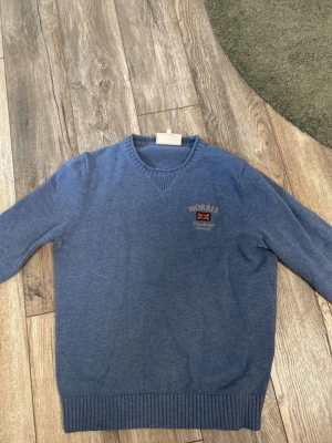 Blå tröja från Morris - Snygg blå tröja från Morris med broderad logga och brittisk flagga på bröstet. Tröjan har V-ringning, ribbade muddar vid ärmslut och nederkant samt är tillverkad i ett mjukt stickat material. Perfekt för en stilren och avslappnad look.