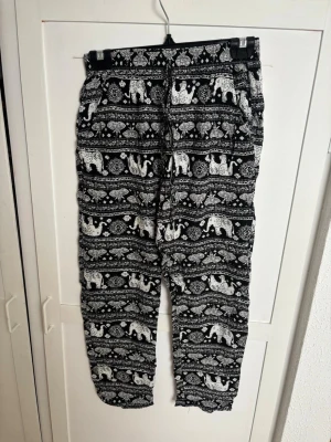 Svarta mjukisbyxor med elefantmönster - Säljer ett par svarta mjukisbyxor med vitt elefantmönster och orientaliska detaljer. Byxorna har resår i midjan och vid bensluten för extra komfort. Perfekta för chill dagar hemma eller när du vill ha en avslappnad stil.