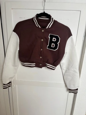 Brun och vit cropped bomberjacka - Snygg brun och vit cropped bomberjacka med stora bokstaven B på bröstet. Jackan har vita ärmar, bruna muddar med vita ränder och tryckknappar framtill. Klassisk collegestil med ribbad krage och muddar. Perfekt för en sportig och trendig look.