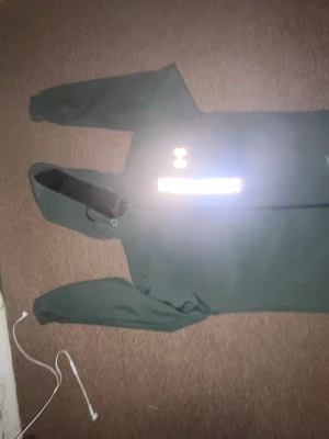 Grön hoodie från Under Armour - Mörkgrön hoodie från Under Armour med dragkedja framtill och stor vit logga samt text på bröstet. Tröjan har huva med dragsko och är tillverkad i ett mjukt, sportigt material som passar perfekt till träning eller chill.