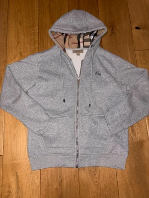 Grå zip hoodie från Burberry - Snygg grå hoodie från Burberry med dragkedja och klassiskt rutigt foder i huvan. Mjuk och skön bomullsblandning, två fickor fram och diskret Burberry-logga på bröstet. Perfekt för en chill och stilren look.