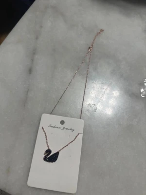 Roséguldhalsband med svart svan - Snyggt halsband i roséguld med en svart svan som hänge. Svanen är dekorerad med små glittrande stenar som ger extra bling. Perfekt accessoar för dig som gillar unika detaljer och djurmotiv.
