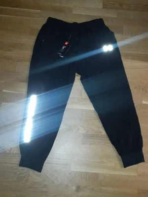 Svarta mjukisbyxor Under Armour - Svarta mjukisbyxor från Under Armour med reflekterande detaljer på ena benet och logga längs sidan. Byxorna har resår i midjan och vid bensluten samt dragsko för bästa passform. Perfekta för chill eller träning.