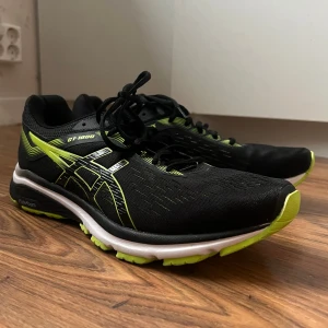 Svarta Asics GT-1000 löparskor - Säljer ett par svarta Asics GT-1000 löparskor med neongröna detaljer och vita inslag på sulan. Skorna har mesh-ovandel för bra ventilation, snörning och en dämpad sula som ger skön komfort vid löpning eller träning. Perfekta för dig som gillar att springa.