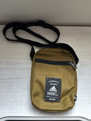 Olivgrön axelväska från Adidas - Snygg olivgrön axelväska från Adidas med svart justerbar axelrem och dragkedja framtill. Väskan har en svart patch med Adidas-logga och text på framsidan. Tillverkad i slitstark textil, perfekt för att bära det viktigaste.