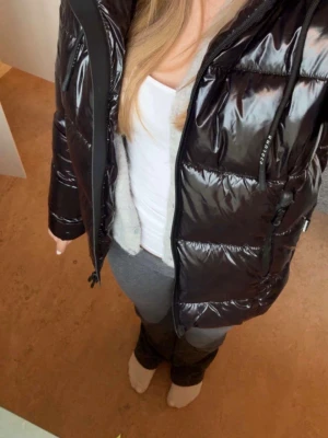 Glansig svart pufferjacka med huva - Snygg svart pufferjacka med glansig finish och huva. Jackan har dragkedja framtill, två sidofickor och quiltad design. Perfekt för kalla dagar och ger en trendig look. Passar dig som vill ha en varm och stilren vinterjacka.