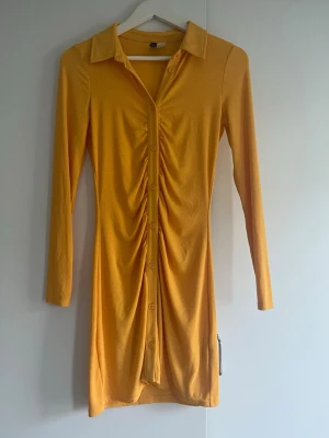 Orange skjortklänning - Trendig gul/orange skjortklänning med långa ärmar och krage. Klänningen har knappar hela vägen framtill och snygg rynkdetalj längs mitten. Figur sydd med stretchigt material!