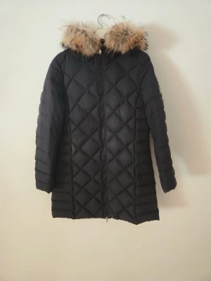 Svart lång dunjacka med fuskpäls - Snygg svart lång dunjacka med quiltat mönster och huva med fluffig fuskpäls. Jackan har dragkedja framtill och är perfekt för kalla vinterdagar. Stilsäker och varm design som håller dig cozy hela säsongen.