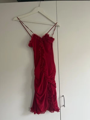 Röd volangklänning med smala axelband - Snygg röd kortklänning med smala axelband och volangdetaljer längs kanterna och framtill. Klänningen har en draperad effekt och är öppen vid brösten. Aldrig använd med lappar kvar.