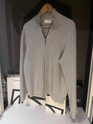 Ljusgrå ziptröja från Selected Homme - Ljusgrå stickad ziptröja från Selected Homme med hög krage och hel dragkedja framtill. Tröjan har ribbade muddar vid ärmslut och nederkant, och är tillverkad i ett mjukt, strukturerat material. Perfekt för lager på lager och enkel att matcha.