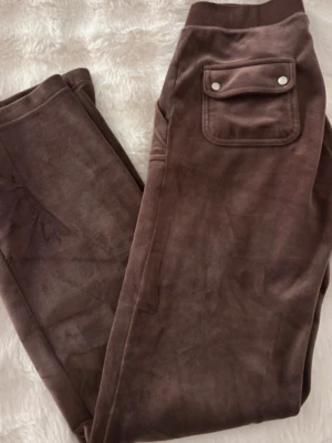 Bruna mjukisbyxor Juicy Couture - Mysiga bruna mjukisbyxor från Juicy Couture i velour med resår i midjan och en ficka bak med knappdetalj. Perfekta för chill dagar och har den klassiska Juicy-looken. Materialet är mjukt och stretchigt för extra komfort.