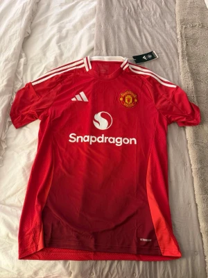 Manchester United matchtröja Adidas - Säljer en röd Manchester United matchtröja från Adidas med vita detaljer och klubbmärke på bröstet. Tröjan har Snapdragons logga på framsidan, klassiska vita ränder på axlarna och är tillverkad i lätt, ventilerande material med Aeroready-teknologi. Helt ny med prislappar kvar och aldrig använd 