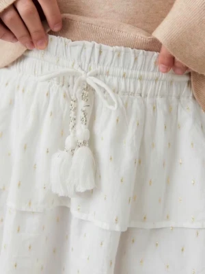 Vit volangkjol med tofsar och snörning - Supersöt vit kortkjol med volanger och små guldiga prickar. Kjolen har snörning i midjan med dekorativa tofsar och pärlor. Perfekt för sommarens alla tillfällen och ger en lätt och luftig känsla. Tillverkad i mjuk bomull.💕