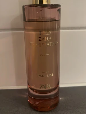 Zara Red Temptation Floral Eau de Parfum - En elegant parfymflaska från Zara med namnet Red Temptation Floral. Flaskan är cylinderformad och har en transparent rosaröd färg med guldfärgad text och lock. Doften är floral och passar dig som gillar fräscha och blommiga parfymer.