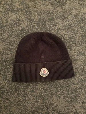 Svart ribbstickad mössa från Moncler - Säljer en svart ribbstickad mössa från Moncler med uppvikt kant och klassisk Moncler-logga framtill. Mössan är tillverkad i mjukt material och har en enkel, stilren design som passar perfekt till kalla dagar.