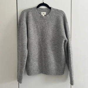 Grå stickad tröja  - Mysig grå stickad tröja med rund hals och ribbade muddar. Tröjan har en klassisk rak passform och är tillverkad i en mjuk ullmix som håller dig varm. Perfekt för lager på lager och enkel att matcha med jeans eller kjol.