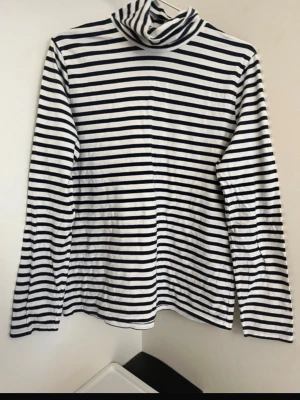 Randig polotröja i svart och vitt - Säljer en långärmad polotröja med klassiska svarta och vita horisontella ränder. Tröjan har en hög krage och är gjord i mjukt, stretchigt material som känns skönt mot huden.