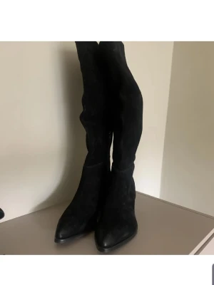 Svarta knähöga mockastövlar - Säljer ett par svarta knähöga stövlar i mocka med spetsig tå och bred klack. Stövlarna har dragkedja på insidan och en stilren design som passar till många outfits. Perfekta för dig som vill ha en edgy och trendig look.