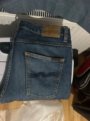 Blå jeans från Nudie Jeans - Säljer ett par klassiska blå jeans från Nudie Jeans med snygg kontrastsöm och ikonisk broderad bakficka. Jeansen har normal passform och är tillverkade i slitstarkt denimtyg. Perfekta för en avslappnad och stilren look.
