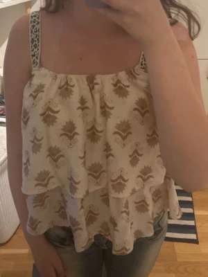 Beige mönstrad linnetopp med volanger - Säljer en luftig linnetopp med breda axelband och volanglager. Köpt i Spanien. Perfekt för varma dagar och ger en avslappnad men trendig look. Kan behövas styrkas lite.