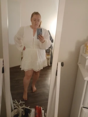 Vit spetsklänning med volang - Säljer en vit klänning med spetsdetaljer och volang nertill. Klänningen har långa, transparenta ärmar och en v-ringning med dekorativa band. Perfekt för dig som gillar romantisk och feminin stil.