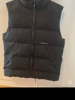 Svart dunväst från Peak Performance - Snygg svart dunväst från Peak Performance med hög krage och dragkedja framtill. Västen har två snedställda fickor och diskret logga på bröstet. Perfekt för lager-på-lager och håller dig varm under kyliga dagar.