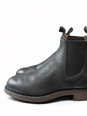 Svarta Chelsea boots i läder - Stilrena svarta Chelsea boots i äkta läder med elastiska sidopaneler och dragflikar både fram och bak. Klassisk rund tå och robust sula i brunt. Perfekta för dig som vill ha en tidlös och enkel look.