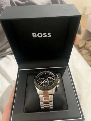 Stilren klocka från BOSS i silver och guld - Snygg klocka från BOSS med svart urtavla och detaljer i guld. Armbandet är i rostfritt stål med inslag av guldtonade länkar. Klockan har flera små urtavlor och en klassisk rund form. Perfekt accessoar för dig som vill ha något elegant och modernt.