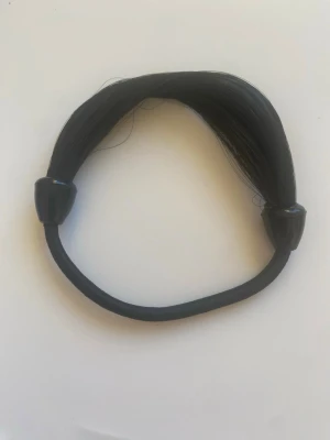 Svart hårsnodd med hår - Cool svart hårsnodd med syntetiskt hår som ger en naturlig look. Perfekt för att snabbt fixa en snygg hästsvans eller knut. Elastisk och enkel att använda, med två svarta plaständar som håller allt på plats.