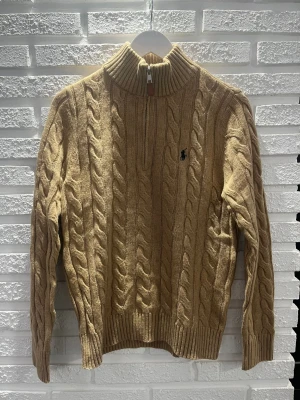 Beige kabelstickad tröja Polo Ralph Lauren - Kabelstickad beige tröja från Polo Ralph Lauren med halv dragkedja och hög krage. Tröjan har det klassiska broderade logotypen på bröstet och ribbade muddar vid ärmslut och nederkant. Perfekt för lager-på-lager och snygg till jeans.