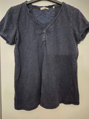 T-shirt - En mörkblå t-shirt i bomull med v-ringning, spetsdetalj vid halsen och tre dekorativa knappar framtill. Kortärmad och normal passform, perfekt för en avslappnad look. Har några hål.