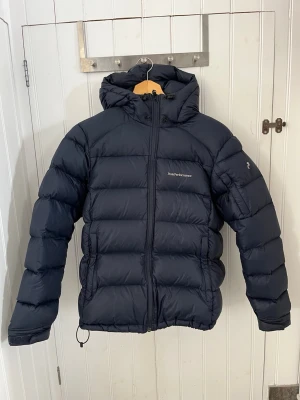 Marinblå dunjacka från Peak Performance - Säljer en marinblå dunjacka från Peak Performance med huva och dragkedja framtill. Jackan har ett puffigt, quiltat mönster och logotyp på bröstet. Perfekt för kalla vinterdagar och tillverkad i slitstarkt material med dunfyllning. Ens spännet där nere är sönder men inget man stör sig på se bild 4