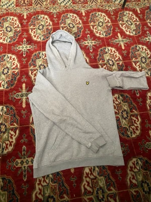 Grå hoodie från Lyle & Scott - Snygg grå hoodie från Lyle & Scott med klassisk gul logga på bröstet. Hoodien har huva med dragsko och en stor magficka. Tillverkad i mjukt bomullsmaterial som känns skönt mot huden. Perfekt för en avslappnad och stilren look.