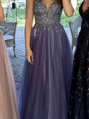 Lång blå balklänning med spetsdetaljer - Elegant långklänning i blått med v-ringad överdel dekorerad med glittriga spetsdetaljer. Kjolen är i flera lager av tunn, svepande tyll som ger en drömmig känsla. Perfekt för bal eller festliga tillfällen. Köpt för 3500kr!!