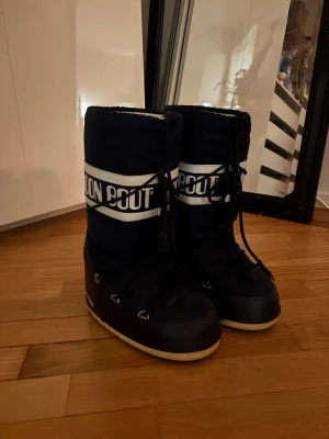 Mörkblå Moon Boot med vit logga - Säljer ett par klassiska mörkblå Moon Boots med vit logga och snörning framtill. De har en hög, vadderad design och vit sula som ger retrovibbar. Perfekta för kalla dagar när du vill sticka ut och hålla värmen.