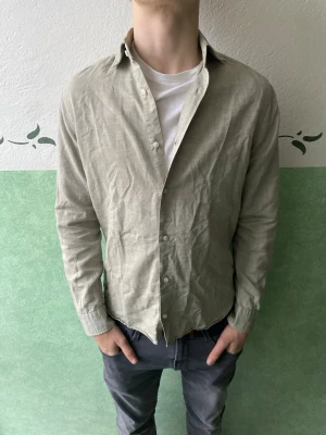 Beige linneskjorta från Riley  - Säljer en stilren beige skjorta från H&M i linne. Skjortan har klassisk krage, vita knappar och långa ärmar med knapp vid ärmslut. Perfekt för dig som gillar en avslappnad och trendig look.