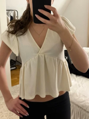 Krämvit med volangärm - Supersöt krämvit topp med v-ringning och volangärmar. Toppen har en lätt ribbad struktur och ett flowy fall nedtill som ger en söt vibe. Perfekt till jeans eller kjol för en trendig look. kontakta för fler bilder på toppen!!