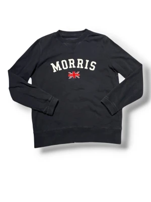 Svart sweatshirt från Morris - Svart sweatshirt från Morris med vit text och en liten brittisk flagga broderad på bröstet. Tröjan har rund halsringning, långa ärmar och ribbade muddar vid ärmslut och nederkant. Perfekt för en avslappnad och stilren look.