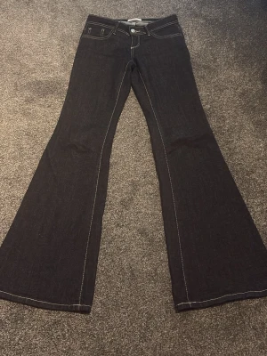 Mörkblå bootcut jeans med kontrastsömmar - Säljer ett par mörkblå bootcut jeans med vita kontrastsömmar och klassiska fem fickor. Jeansen har hög midja och vida ben, vilket ger en retroinspirerad look. Materialet är kraftig denim som känns slitstark och snygg.