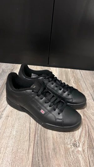 Svarta Reebok Classic sneakers - Stilrena svarta Reebok Classic sneakers i lågt snitt med ovandel i slätt läder och matchande svarta skosnören. Diskret Reebok-logga och brittiska flaggan på sidan. Perfekta för dig som gillar en clean och tidlös look.