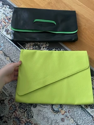 Neongul kuvertväska i konstläder - Säljer två snygga neongul och svart/grön kuvertväskor i konstläder med stilren, asymmetrisk flärp. Väskor har en slät yta och är perfekt för dig som vill sticka ut. Enkel och modern design utan synligt märke. Passar till dig som gillar färg och trendiga accessoarer. Säljer båda tillsammans! 