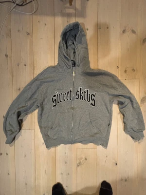 Grå Sweet Sktbs zip hoodie - Grå hoodie med dragkedja från Sweet Sktbs. Stor huva och två fickor framtill. Tryck i svart gotisk stil på bröstet. Tillverkad i mjukt bomullsmaterial, perfekt för en avslappnad streetwear-look. Säljer då jag växte ur den tätt inpå att jag köpte den.
