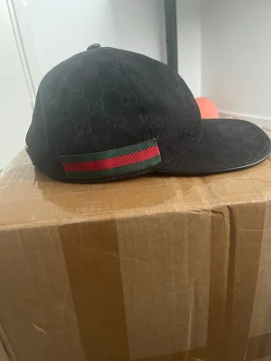 Svart keps från Gucci med GG-mönster - Svart keps från Gucci med klassiskt GG-monogram över hela tyget och en ikonisk grön-röd rand på sidan. Kepsen har böjd skärm och är tillverkad i canvasmaterial. Perfekt för dig som vill ha en stilren och lyxig accessoar. Kan mötas i jkpg vmo gvd och mer