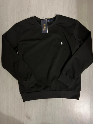 Svart sweatshirt från Polo Ralph Lauren - Klassisk svart sweatshirt från Polo Ralph Lauren med rund halsringning och diskret vit logga broderad på bröstet. Tröjan har ribbade muddar vid ärmslut och nederkant, perfekt för en clean och stilren look.