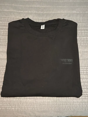 Svart Moschino sweatshirt med logga - Säljer en svart sweatshirt från Moschino med vita och röda band längs ärmarna där det står 'Moschino' i svart text. Tröjan har rund halsringning och ribbade muddar vid ärmslut och nederkant. Materialet är mjukt och bekvämt, perfekt för en trendig streetwear-look.