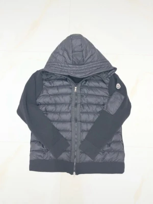 Svart pufferjacka från Moncler - Snygg svart pufferjacka från Moncler med quiltad kropp och stickade ärmar. Jackan har huva, dragkedja framtill och en ficka med dragkedja på ärmen där Moncler-loggan sitter. Perfekt för kalla dagar och riktigt trendig look.