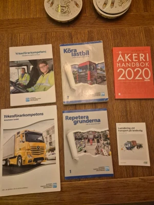 Kurslitteratur för lastbilsförare - Samling kurslitteratur för lastbilsförare, inklusive böcker som 'Yrkesförarkompetens', 'Köra lastbil', 'Åkeri Handbok 2020', 'Repeterar grunderna' och 'Lastsäkring vid transport på landsväg'. Böckerna har färgglada omslag med bilder på lastbilar och transportmiljöer.