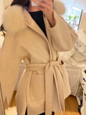 Beige kappa med pälskrage och knytskärp - Stilren beige kappa med pälskrage och knytskärp i midjan. Kappan har en rak passform, vida ärmar och är tillverkad i ett mjukt material som ger en lyxig känsla. Passar S-M