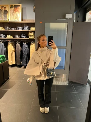 Beige poncho - Snygg beige poncho kappa med hög krage och knytband i midjan. Kappan har vida ärmar och en dekorativ knapp framtill. Perfekt för att ge en elegant touch till din outfit. Från missay passar perfekt till vår/sommar. Har en fläck men går säkert att få bort.