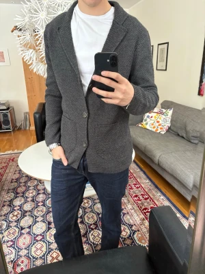 Mörkgrå stickad cardigan - Snygg mörkgrå stickad cardigan med 3 knappar framtill och två stora fickor. Cardiganen har en avslappnad passform och mjukt material som ger en casual men ändå stilren look. 